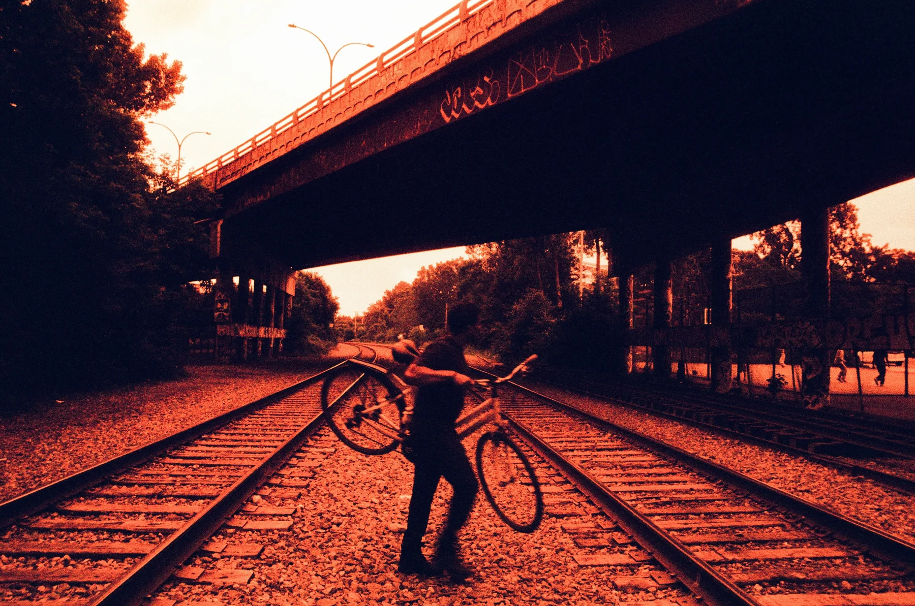 A person with a bicycle on railway tracks beneath an overpass, in red and orange tones